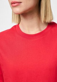 Rød bomulds t-shirt med rund hals, korte ærmer og en glat tekstur. Har syninger langs halsudskæringen og skuldersømmene.