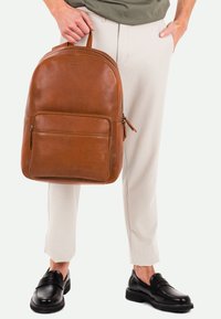 Mochila de cuero marrón con bordes redondeados, bolsillo frontal con cremallera y asa superior. Lleva puesta con pantalones de color claro y zapatos negros.