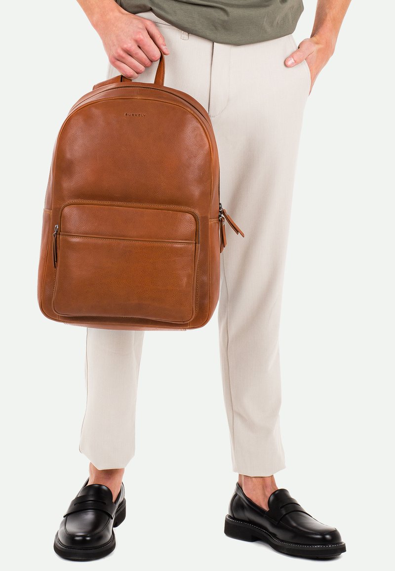 Mochila de cuero marrón con bordes redondeados, bolsillo frontal con cremallera y asa superior. Lleva puesta con pantalones de color claro y zapatos negros.