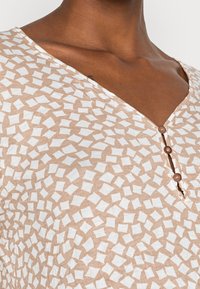 Tunika beige con un motivo geometrico bianco testurizzato, scollatura a V e tre bottoni marroni sul davanti. Tessuto liscio e leggero.