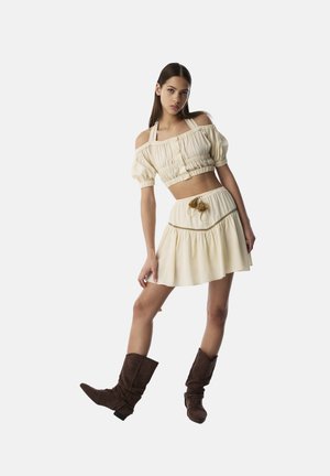 Top court crème à épaules dénudées avec des manches bouffantes et une jupe évasée assortie, dotée d'un cordon de serrage décoratif et d'un éclat subtil. Bottes marron.