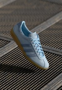 Sapatilha de camurça azul clara com sola de borracha e cordões combinando, repousando sobre um fundo de grade metálica industrial.