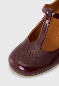 Burgundy leather shoe featuring a rounded toe, T-strap design, perforated detailing, and a smooth, shiny finish.