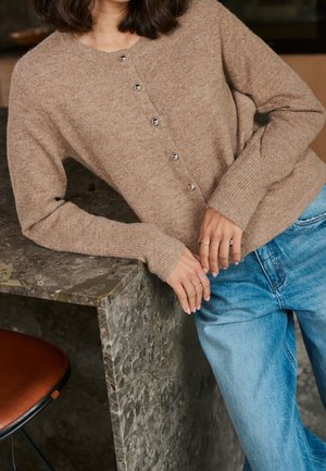 Personne portant un cardigan marron boutonné et un jean bleu, appuyée sur une table en marbre gris, les mains reposant sur le bord.