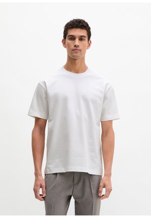 Młody mężczyzna w prostej, białej koszulce z krótkim rękawem i szarych spodniach, stojący na jednolitym białym tle.