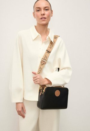 Kvinde iført hvid sweater med krave og bukser, der holder en sort, kasseformet taske med en brun justerbar rem med trykt mærkenavn.