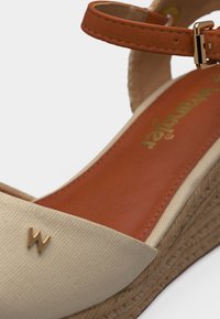 Μπεζ πλατφόρμα σανδάλι με πλεκτή σόλα από γιούτα, καφέ δερμάτινο λουράκι αστραγάλου με χρυσή αγκράφα και μικρό χρυσό έμβλημα "W" στην περιοχή των δαχτύλων.