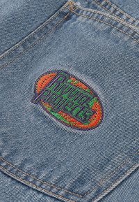 Blåt denimstof med en broderet oval lap, der har teksten "Running Projects" i grønt og lilla, omgivet af en orange kant.