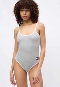 Γυναίκα που φοράει γκρι ριμπ ολόσωμο κορμάκι με λεπτές τιράντες και μικρό λογότυπο Tommy Hilfiger κοντά στο ισχίο, στέκεται μπροστά σε απλό φόντο.