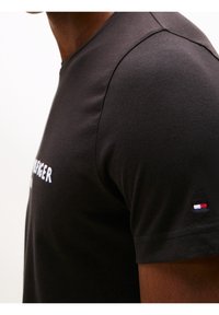 Czarny bawełniany t-shirt z okrągłym dekoltem, z białym logo i małym akcentem flagi w kolorach czerwonym, białym i niebieskim na rękawie.