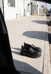 Paire de mocassins noirs à semelle épaisse posée sur un trottoir vide à côté d’une porte de voiture dans une zone urbaine avec des bâtiments blancs.