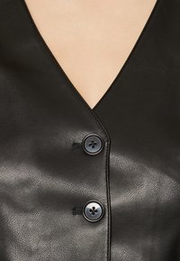 Czarna skórzana odzież z głębokim dekoltem V oraz dwoma okrągłymi czarnymi guzikami. Tekstura jest gładka z subtelnym ziarnem.