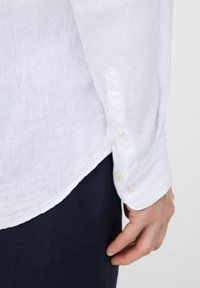 Camicia bianca di lino con maniche lunghe, dotata di chiusura con bottone ai polsi e orlo leggermente curvato. Tessuto liscio e materiale leggero.
