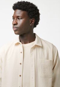 Beige button-down overhemd in een gestructureerde stof, met een borstzak en een losse pasvorm, gedragen over een bijpassend lichtkleurig t-shirt.