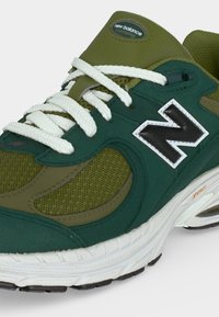 Πράσινο και ελιά New Balance παπούτσι για τρέξιμο με λευκά κορδόνια, μαύρο και λευκό λογότυπο και λευκή σόλα με λεπτομέρειες αποσβεσης.