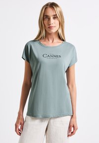 Světle zelené tričko s krátkým rukávem a kulatým výstřihem, s černým grafickým textem "Cannes". Měkká tkanina a volný střih.