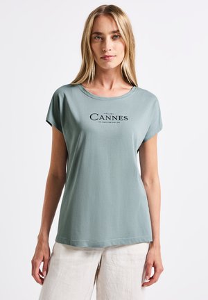 Helles grünes T-Shirt mit kurzen Ärmeln und rundem Halsausschnitt, mit einem schwarzen "Cannes"-Grafiktext. Weicher Stoff und lockere Passform.