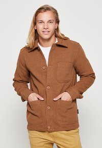Brown herringbone-patterned jacket with a collared neckline, long sleeves, two front pockets, and button closures, worn over a white shirt.