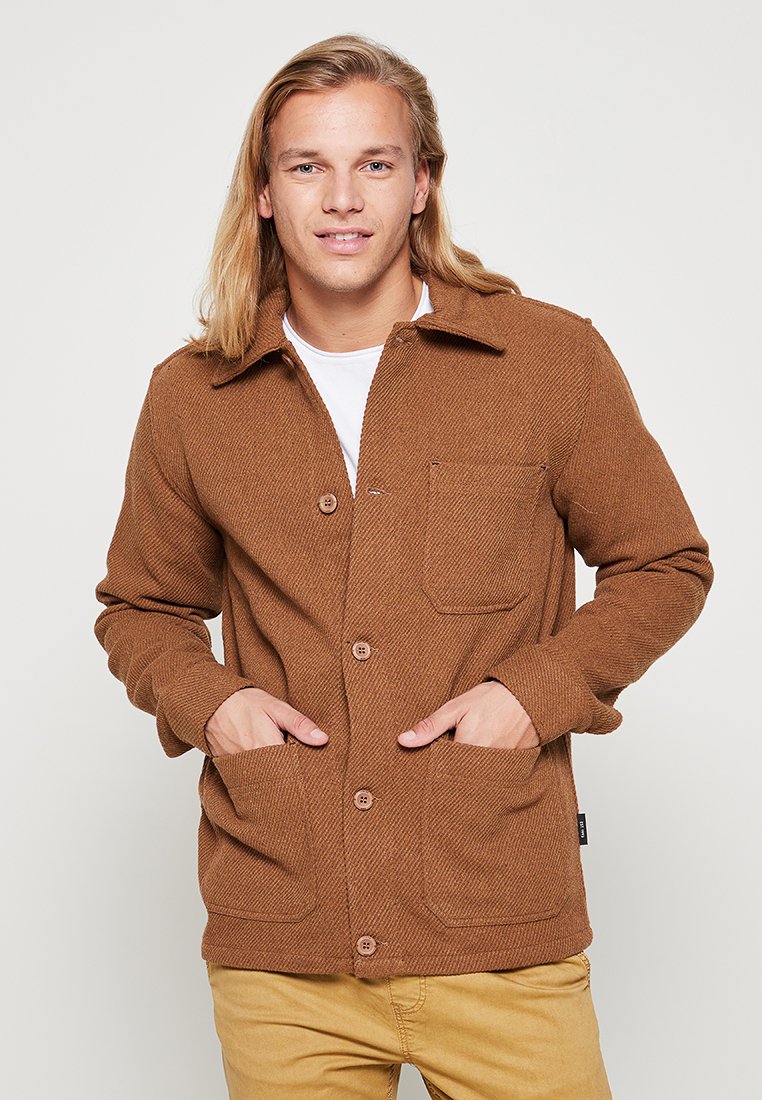 Brown herringbone-patterned jacket with a collared neckline, long sleeves, two front pockets, and button closures, worn over a white shirt.