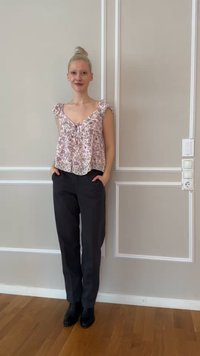 Νεαρό άτομο με ξανθά μαλλιά σε κότσο, φορώντας ροζ μπλούζα με floral και κοντά μανίκια, μαύρο παντελόνι και μαύρα παπούτσια, στέκεται μπροστά σε ανοιχτό μπεζ τοίχο.