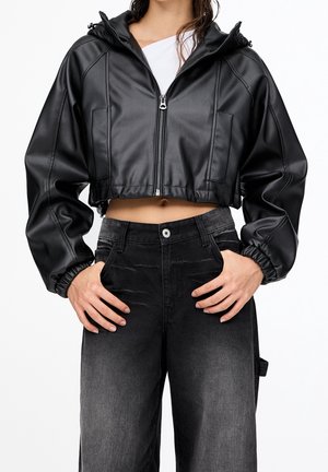 Kvinde iført en kort sort faux læderjakke med lynlås og elastiske manchetter samt højtaljede, sortfasede jeans, hænderne hvilende på hofterne.