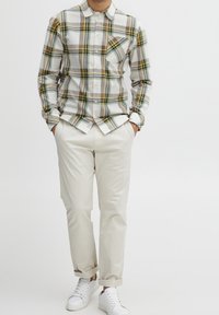 Mann in kariertem, weiß-gelben Hemd, beigen, hochgekrempelten Hosen, mit den Händen in den Taschen und weißen Turnschuhen vor einfarbigem Hintergrund.