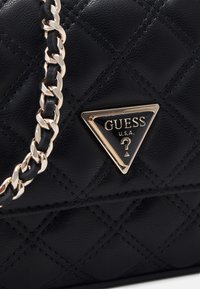 Musta, tikattu käsilaukku, valmistettu keinonahasta. Varustettu kultaisen sävyisellä ketjuhihnalla ja kolmionmuotoisella logoetiketillä, jossa lukee "GUESS".