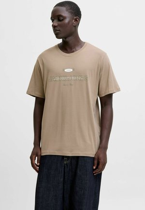 Vīrietis, tērpies bēša krāsas īsām piedurknēm T-krekliņā ar uzrakstu "1990 Jack & Jones" un tumšās platās biksēs, stāv uz vienkārša baltā fona.