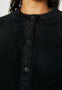 Zwarte, pluizige cardigan met een ronde hals, zichtbare stiksels en zwarte knopen aan de voorkant om te sluiten. Zachte textuur.