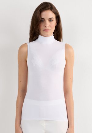 Ung kvinde med langt brunt hår iført en tætsiddende hvid ærmeløs rullekrave top mod en ensfarvet lys baggrund.