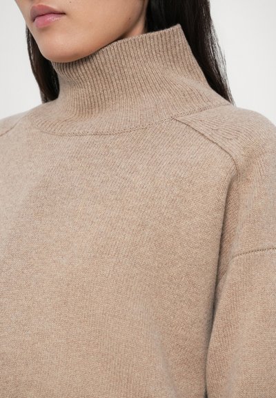 Vrouw in een beige geribde turtleneck trui, waarbij de textuur en pasvorm rond de nek en schouders zichtbaar zijn.
