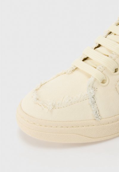 Canvas sneakers i cremefarve med frynset kanter, rund tå, snørebåndsdesign og struktureret gummisål. Nærbillede, der fremhæver detaljerne.