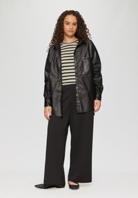 Zwarte faux leren shirtjas met twee borstzakken, gelaagd over een gestreepte lange mouwen top. Wijde zwarte broek en platte zwarte schoenen.
