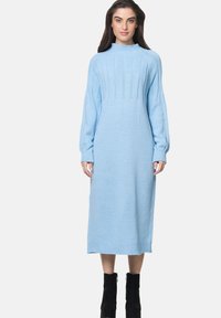 Abito maglione a maglia di colore blu chiaro con collo alto, maniche lunghe, dettagli a costine e taglio medio. Indossato con stivaletti neri alla caviglia.