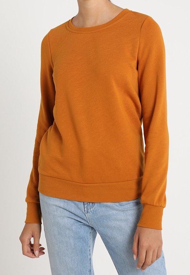 Femme portant un pull orange uni à manches longues et un jean bleu clair, debout devant un fond blanc uni.