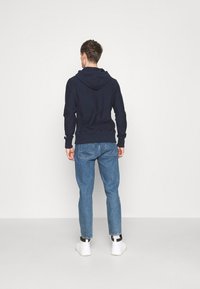Sweat à capuche navy avec cordon de serrage, associé à un jean bleu clair et des baskets blanches. La texture semble douce ; le design présente une silhouette simple.
