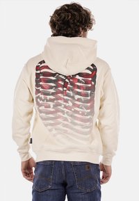 Felpa con cappuccio color crema con una grande grafica di un design in rosso e nero che riproduce una gabbia toracica sul retro. Tessuto morbido con una vestibilità rilassata.