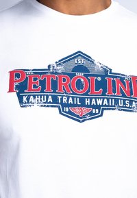 Camiseta de algodón blanca con un gráfico de estilo vintage en azul y rojo, que muestra "PETROL INN" con un emblema alado y el año 1989.