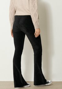 Persona che indossa pantaloni a zampa d'elefante in corduroy neri, un maglione beige a trecce e sneakers bianco e nere, in piedi contro un muro bianco uniforme.