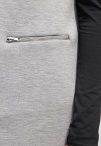 Szary materiał o miękkiej fakturze z kieszenią zapinaną na suwak. Projekt obejmuje czarny rękaw kontrastujący z szarym ciałem. Widoczne elementy metalowe suwaka.