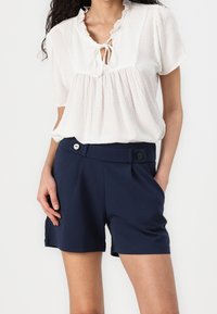 Femme portant un chemisier blanc texturé à manches courtes avec un col tie et un short bleu marine ajusté avec des détails de boutons, les mains dans les poches.
