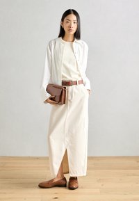 Camisa blanca de botones sobre una blusa beige, falda crema con abertura lateral, cinturón de cuero marrón y mocasines marrones; llevando un bolso de mano de cuero marrón texturizado.