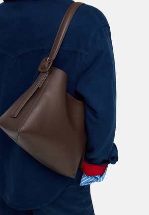 Osoba ubrana w ciemnoniebieską kurtkę i czerwoną rękawicę swetra, nosząca na ramieniu dużą brązową skórzaną torbę na ramię.