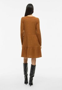 Robe marron à manches longues côtelées avec jupe à volants, dotée d'un col rond classique. Portée avec des bottes hautes noires. Texture lisse.