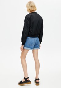 Personne blonde portant une veste bomber noire, un short en jean retroussé et des sandales à plateforme noires, debout de dos sur un fond blanc.
