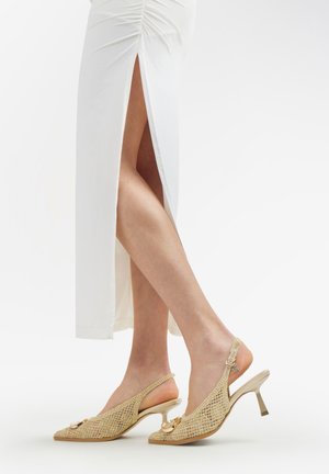 Gambe in gonna bianca con spacco laterale che indossano décolleté slingback beige intrecciate con tacco sottile di altezza media e dettaglio catena color oro.
