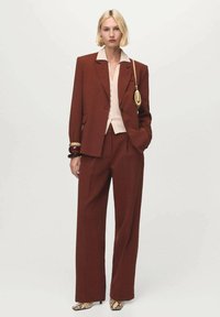 Mujer con traje entallado marrón óxido, blusa crema, tacones con estampado de serpiente, llevando un pequeño bolso dorado, de pie contra un fondo liso.