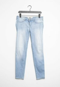 Ljust blå denimjeans med slim fit, blekt tvätt, fem fickor och knappstängning, hängande på en svart galge mot en vit bakgrund.