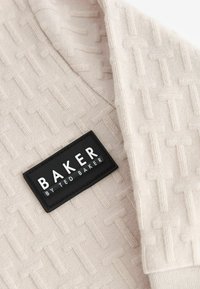 Béžová textúrovaná látka s opakujúcim sa vzorom. Čierna etiketa s nápisom "BAKER" a "by TED BAKER" v bielom písmene.