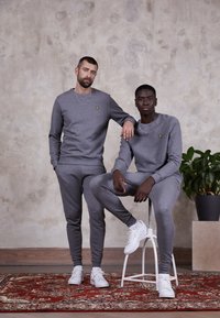 Un sweat-shirt gris et un jogging, avec un petit logo jaune. Les deux modèles portent des baskets blanches ; en arrière-plan, il y a une plante en pot et un tapis à motifs.